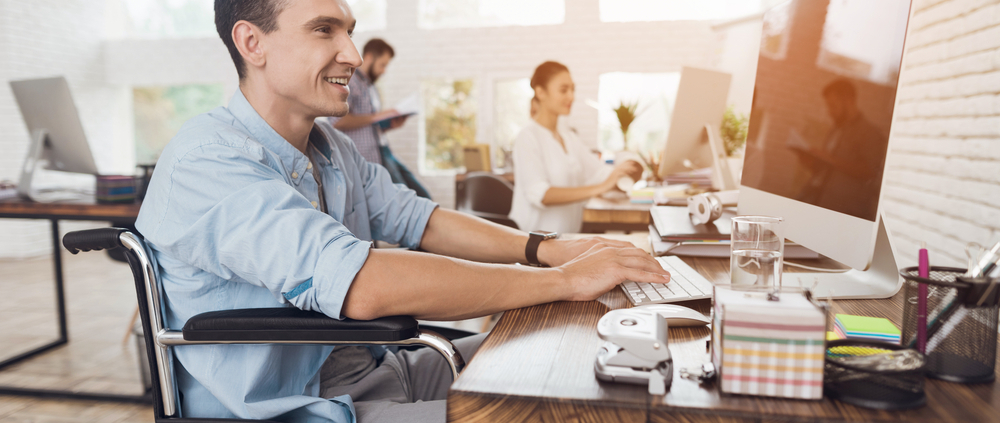Ein fröhlicher Mann im Rollstuhl sitzt an in einem Büro am Computer. Im Hintergrund sind Kolleg:innen zu sehen
