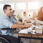 Ein fröhlicher Mann im Rollstuhl sitzt an in einem Büro am Computer. Im Hintergrund sind Kolleg:innen zu sehen