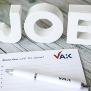 Das Wort Job in weißen Buchstaben auf grauem Holzhintergrund, davor liegend ein VAK-Schreibblock uund ein VAK-Kugelschreiber