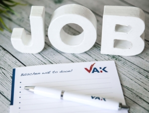 Job Das Wort Job in weißen Buchstaben auf grauem Holzhintergrund, davor liegend ein VAK-Schreibblock uund ein VAK-Kugelschreiber