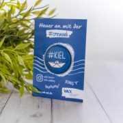 blaues Giveaway mit Beschriftung "Heuer an" zum Thema Jobsuche auf einem grauen Holzhintergrund mit einer Grünpflanze im Hintergrund