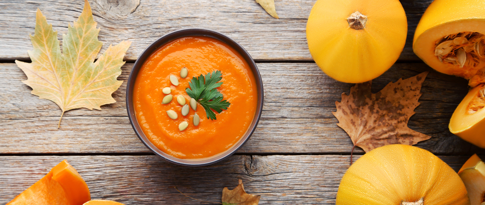 Eine Schale Kürbissuppe steht auf einem hellen Holzuntergrund, daneben liegen verschiedene Kürbisse