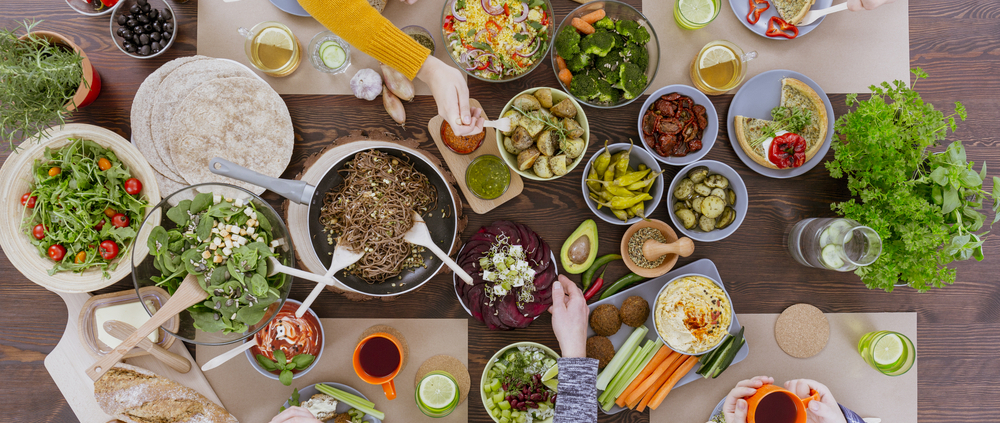 Ansicht von oben auf einen Tisch mit Schüsseln gefüllt mit verschiedenen Lebensmitteln wie Salat, Antipasti, Gemüse; vier Personen sitzen am Tisch, zu sehen sind ihre Arme und Hände.