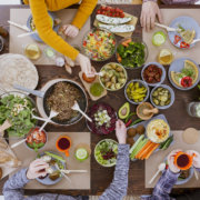 Ansicht von oben auf einen Tisch mit Schüsseln gefüllt mit verschiedenen Lebensmitteln wie Salat, Antipasti, Gemüse; vier Personen sitzen am Tisch, zu sehen sind ihre Arme und Hände.