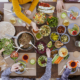 Ansicht von oben auf einen Tisch mit Schüsseln gefüllt mit verschiedenen Lebensmitteln wie Salat, Antipasti, Gemüse; vier Personen sitzen am Tisch, zu sehen sind ihre Arme und Hände.