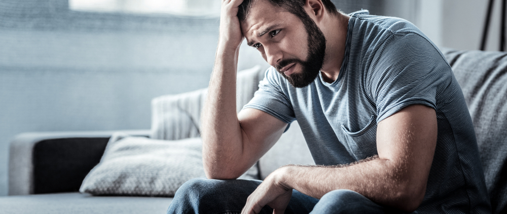 Ein dunkelhaariger Mann sitzt und schaut deprimiert, eine Hand hält er sich an den Kopf